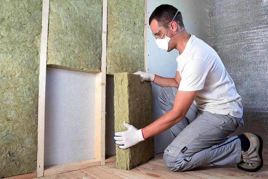 Worker in protective goggles and respirator insulating rock wool insulation in wooden frame for future house walls for cold barrier. Comfortable warm home, economy, construction and renovation concept