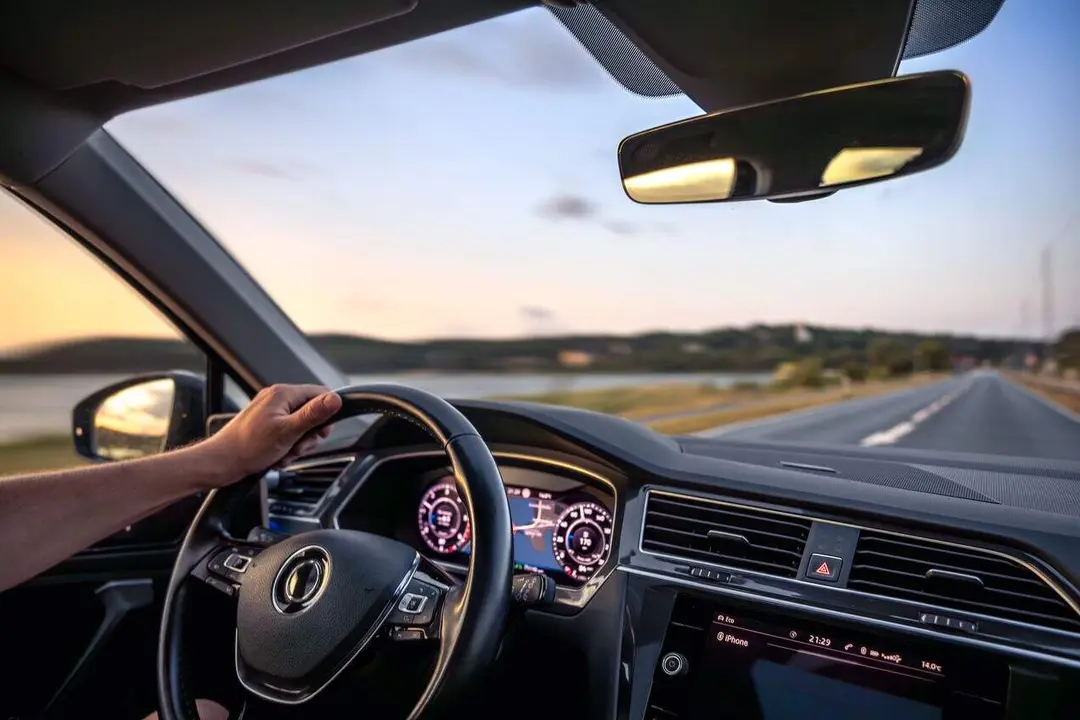 A driver's hand on the wheel during a peaceful sunset drive along a riverside highway, with dashboard navigation and glowing ambient light.