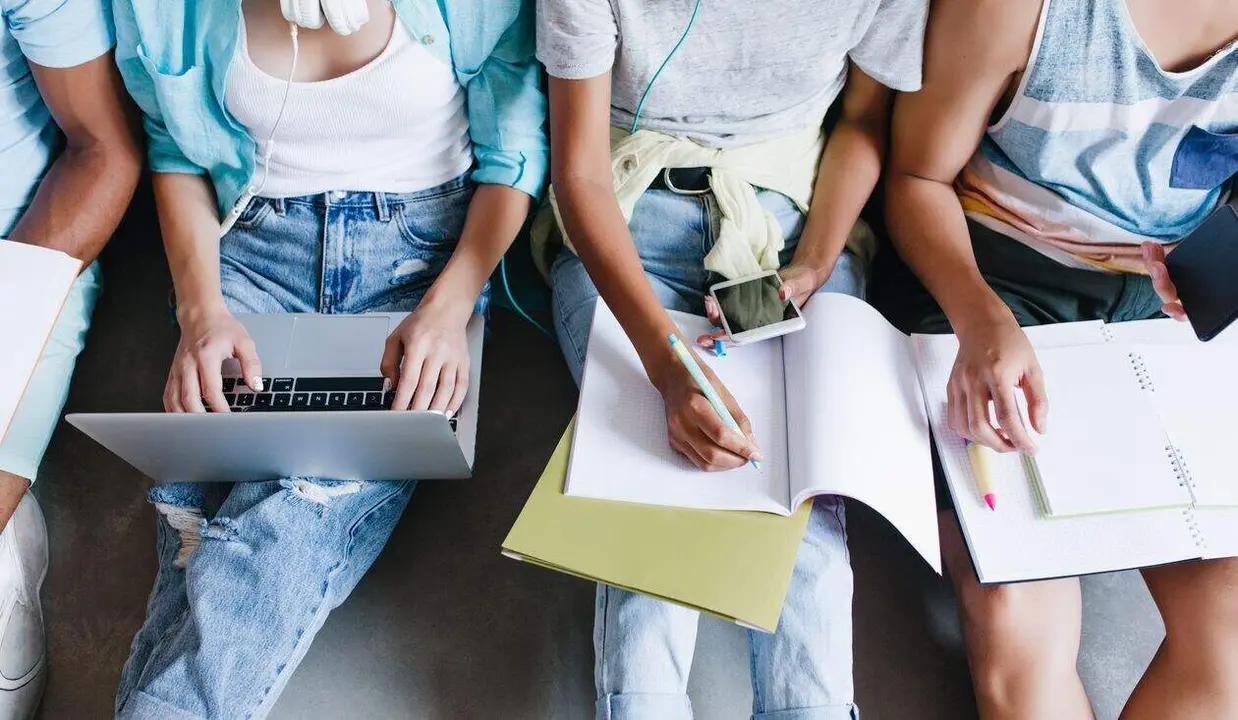 Close-up overhead portrait of girl in blue shirt and jeans holding laptop on knees while sitting beside university mates. Female student writing lecture in notebook and using phone between friends.