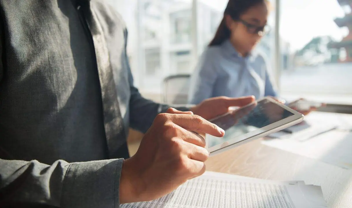 Unrecognizable Asian businessman wearing grey shirt sitting at table in office and browsing information on touchpad. Documents lying on table, female colleague working in background