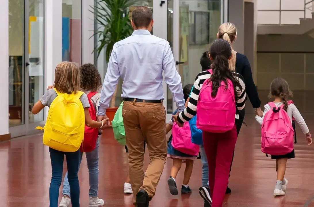 Schoolkids with bright backpacks walking through school hallway, holding hands of teachers. Back view, full length. Education or back to school concept