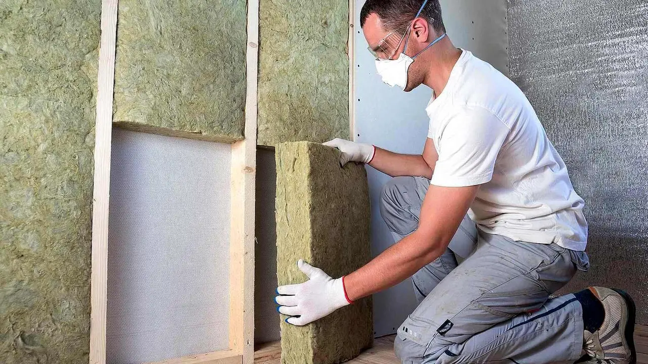 Worker in protective goggles and respirator insulating rock wool insulation in wooden frame for future house walls for cold barrier. Comfortable warm home, economy, construction and renovation concept