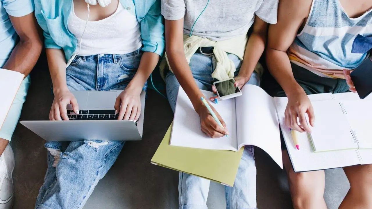 Close-up overhead portrait of girl in blue shirt and jeans holding laptop on knees while sitting beside university mates. Female student writing lecture in notebook and using phone between friends.