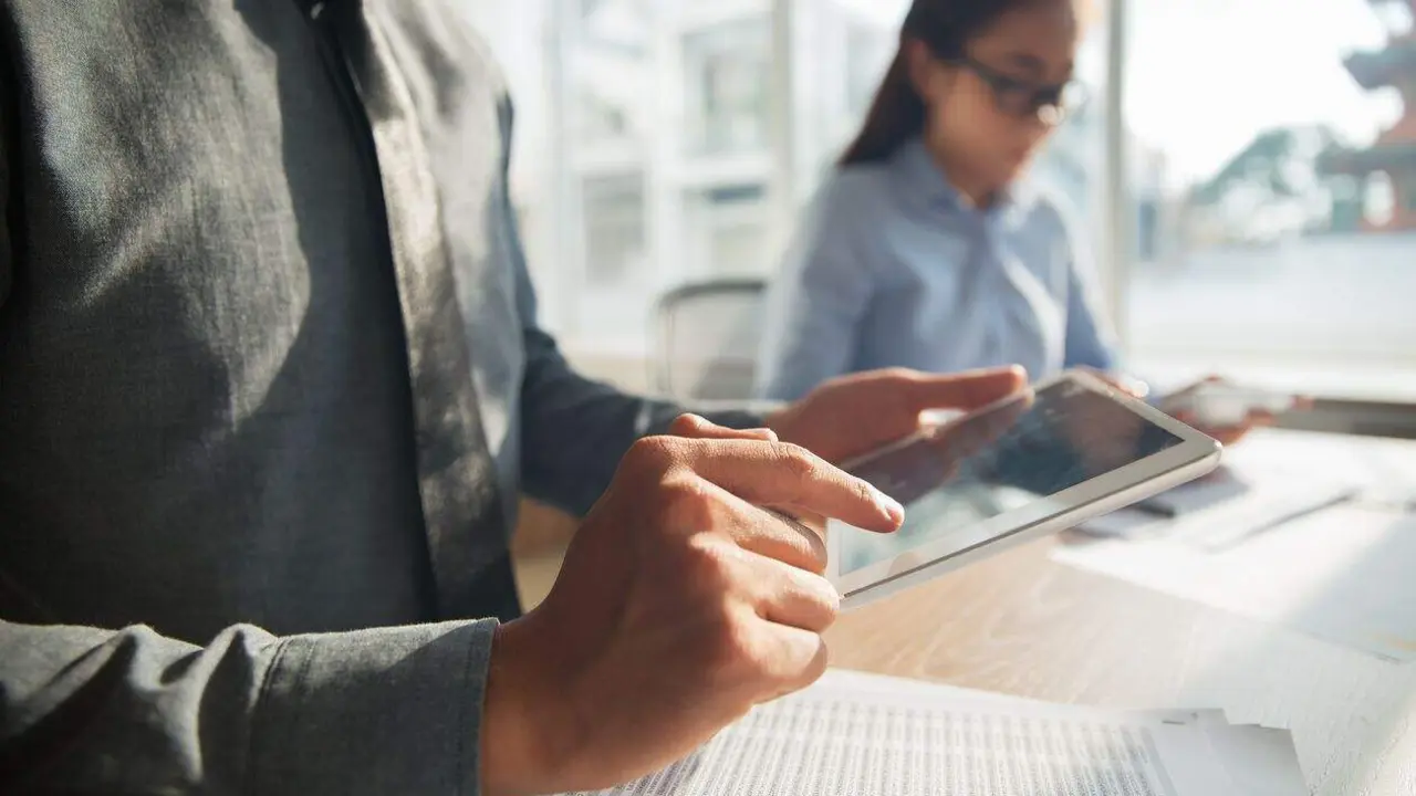 Unrecognizable Asian businessman wearing grey shirt sitting at table in office and browsing information on touchpad. Documents lying on table, female colleague working in background