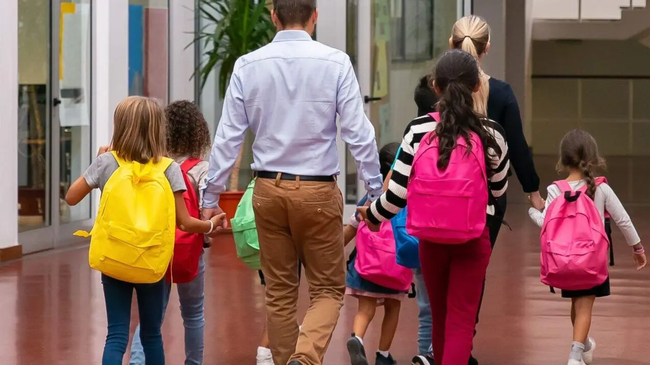 Schoolkids with bright backpacks walking through school hallway, holding hands of teachers. Back view, full length. Education or back to school concept
