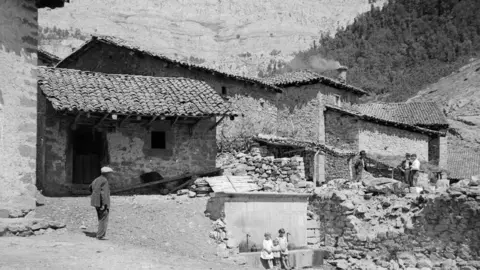 Eslatal gegen Riaño. Salas & Peñas Las Pintas [Valle del Esla en los alrededores de Riaño. Salas en primer plano y peñas Pintas al fondo. Niñas apoyadas en la fuente, un hombre a la izquierda caminando y tres hombres conversando a la derecha]